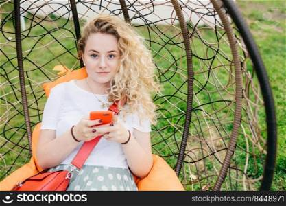 People and leisure concept. Beautiful curly female holds smart phone, types text message, looks at camera, has good rest in hanging chair outdoor, dressed in casual clothes, has appealing look