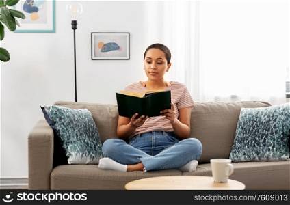 people and leisure concept - happy african american woman sitting on sofa and reading book at home. african american woman reading book at home