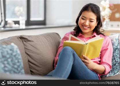 people and leisure concept - happy asian young woman in pink shirt sitting on sofa and reading book at home. asian young woman reading book at home