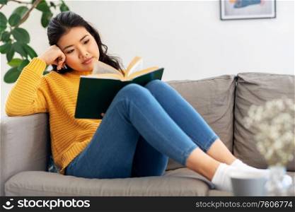 people and leisure concept - happy asian young woman in yellow sweater sitting on sofa and reading book at home. asian young woman reading book at home