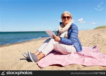 people and leisure concept - happy senior woman writing to notebook on summer beach in estonia. senior woman writing to notebook on summer beach