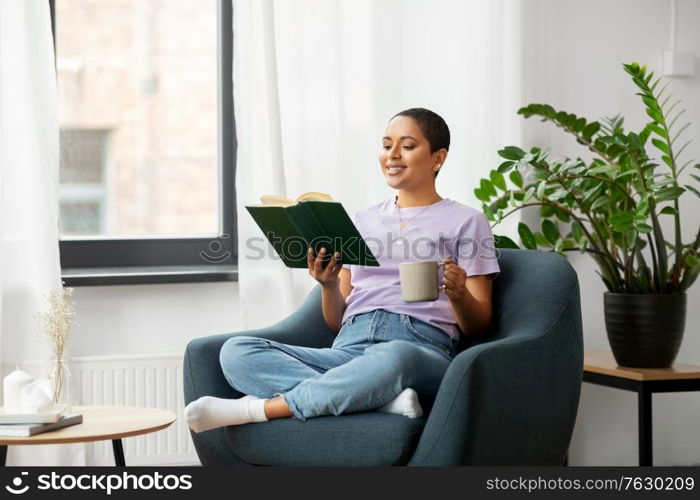 people and leisure concept - happy smiling african american woman in glasses sitting on sofa and reading book at home. happy african american woman reading book at home