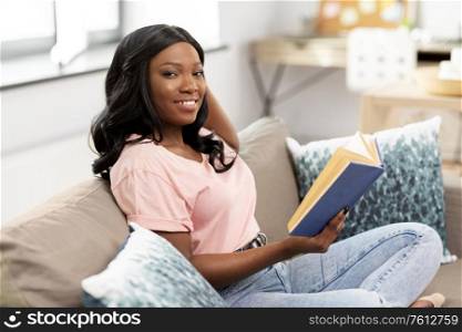 people and leisure concept - happy smiling african american woman sitting on sofa and reading book at home. happy african american woman reading book at home