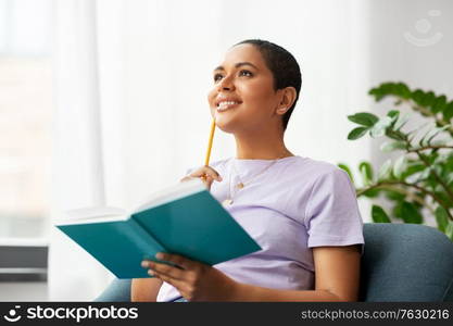 people and leisure concept - happy smiling african american woman with diary and pencil sitting in chair at home. happy african american woman with diary at home