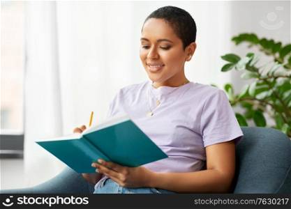 people and leisure concept - happy smiling african american woman with diary sitting in chair at home. happy african american woman with diary at home