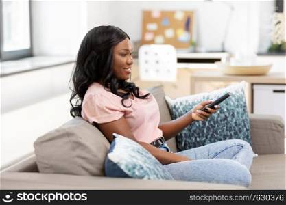 people and leisure concept - happy smiling african american young woman with remote control watching tv at home. happy african american woman watching tv at home