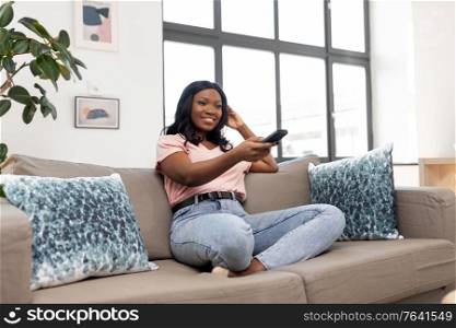 people and leisure concept - happy smiling african american young woman with remote control watching tv at home. happy african american woman watching tv at home