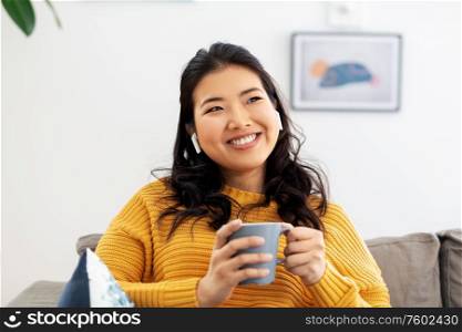 people and leisure concept - happy smiling asian young woman in earphones listening to music and drinking coffee at home. woman in earphones listening to music at home
