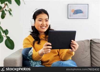 people and leisure concept - happy smiling asian young woman in headphones listening to music on tablet pc computer at home. asian woman with headphones and tablet pc at home