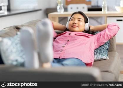 people and leisure concept - happy smiling asian young woman in headphones listening to music lying on sofa at home. woman in headphones listening to music at home