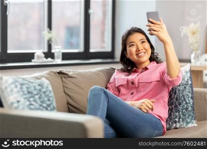 people and leisure concept - happy smiling asian young woman in pink shirt sitting on sofa and taking selfie with smartphone at home. asian woman taking selfie with smartphone at home