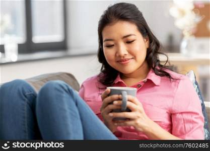 people and leisure concept - happy smiling asian young woman in pink shirt sitting on sofa and drinking coffee at home. smiling asian young woman drinking coffee at home