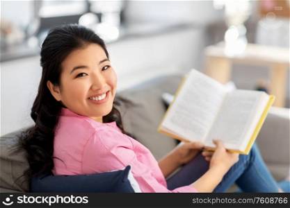 people and leisure concept - happy smiling asian young woman in pink shirt sitting on sofa and reading book at home. smiling asian young woman reading book at home