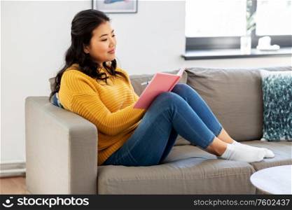 people and leisure concept - happy smiling asian young woman in yellow sweater with diary or notebook sitting on sofa at home. asian woman with diary sitting on sofa at home