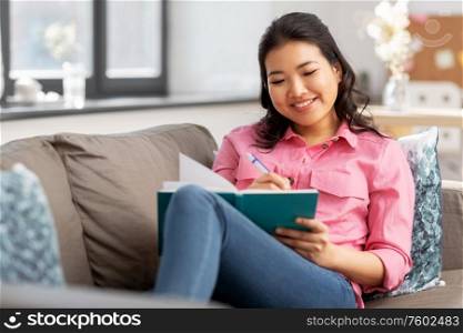 people and leisure concept - happy smiling asian young woman with diary or notebook sitting on sofa at home. asian woman with diary sitting on sofa at home