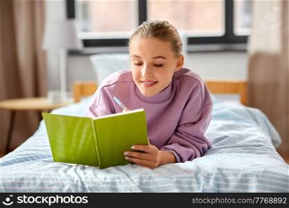 people and leisure concept - happy smiling girl writing to diary lying on bed at home. smiling girl writing to diary lying on bed at home