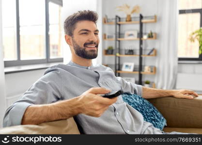 people and leisure concept - happy smiling man with remote control watching tv at home. happy man with remote control watching tv at home