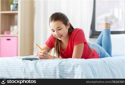 people and leisure concept - happy smiling teenage girl with diary and pencil lying on bed at home. teenage girl writing to diary at home