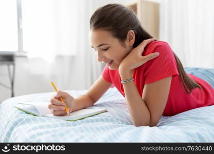 people and leisure concept - happy smiling teenage girl with diary and pencil lying on bed at home. teenage girl writing to diary at home