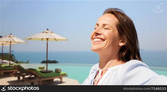 people and leisure concept - happy smiling woman enjoying sun over infinity edge pool of touristic resort background. happy woman enjoying sun over infinity edge pool