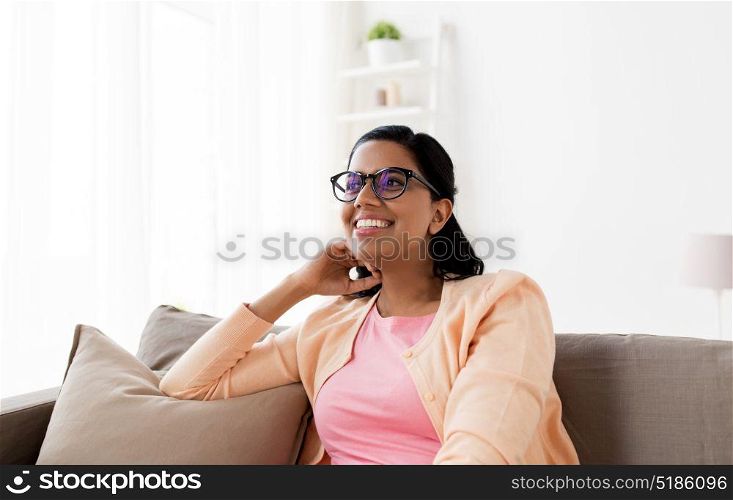 people and leisure concept - happy smiling young indian woman in glasses sitting on sofa at home. happy smiling young woman sitting on sofa at home
