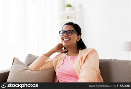 people and leisure concept - happy smiling young indian woman in glasses sitting on sofa at home. happy smiling young woman sitting on sofa at home