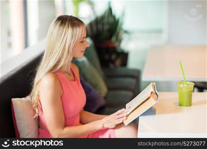 people and leisure concept - happy woman with green smoothie drink or vegetarian shake reading book at restaurant or cafe. woman with drink reading book at cafe