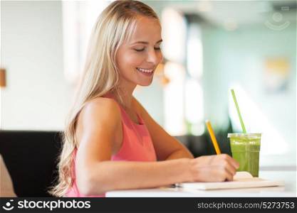 people and leisure concept - happy woman with green smoothie drink or vegetarian shake writing to notebook at restaurant or cafe. woman with drink writing to notebook at restaurant
