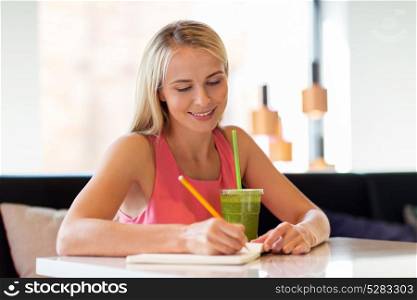 people and leisure concept - happy woman with green smoothie drink or vegetarian shake writing to notebook at restaurant or cafe. woman with drink writing to notebook at restaurant