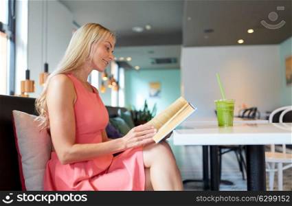 people and leisure concept - happy woman with green smoothie drink or vegetarian shake reading book at restaurant or cafe. woman with drink reading book at cafe