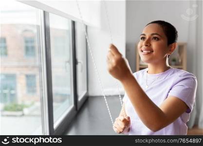 people and leisure concept - happy young african american woman opening window roller blinds at home. woman opening window roller blinds