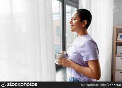 people and leisure concept - happy young african american woman with coffee cup looking out of window at home. happy woman with coffee looking out of window