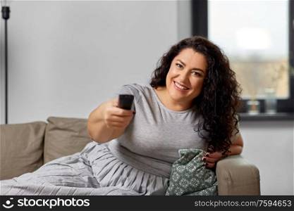 people and leisure concept - happy young woman lying on sofa with remote control and watching tv at home. woman with remote control and watching tv at home