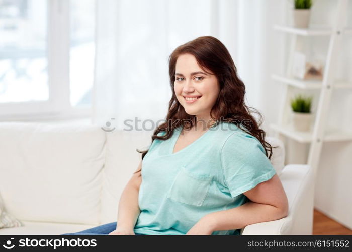 people and leisure concept - happy young woman plus size sitting on sofa at home. happy young plus size woman at home