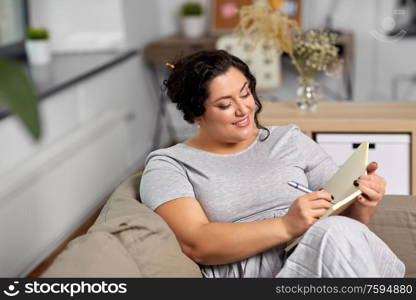 people and leisure concept - happy young woman with diary and pen taking notes on sofa at home. happy young woman with diary on sofa at home