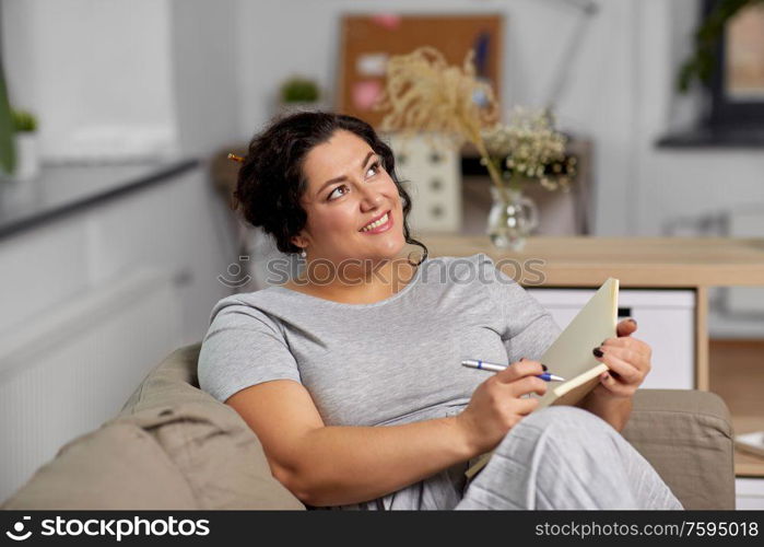 people and leisure concept - happy young woman with diary and pen taking notes on sofa at home. happy young woman with diary on sofa at home