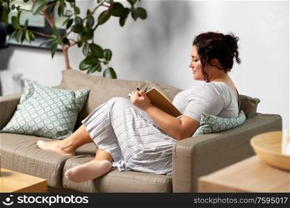 people and leisure concept - happy young woman with diary and pencil taking notes on sofa at home. happy young woman with diary on sofa at home
