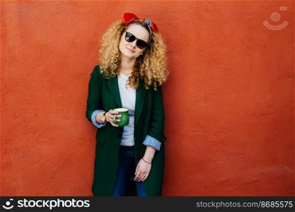 People and leisure concept. Horizontal portrait of pretty girl wearing headband, jacket, jeans and sunglasses holding cup of hot drink, having tea or coffee, posing against orange background