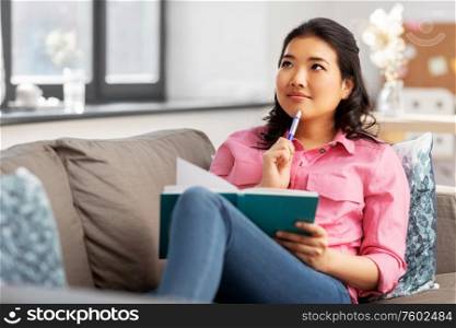people and leisure concept - thinking asian young woman with diary or notebook sitting on sofa at home. asian woman with diary sitting on sofa at home