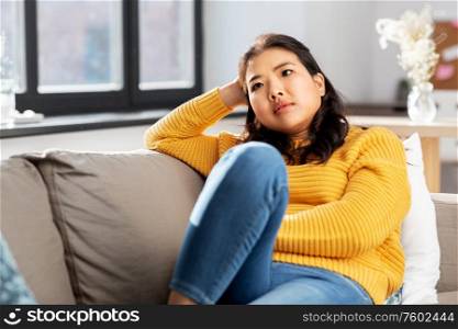 people and leisure concept - worried asian young woman in yellow sweater sitting on sofa at home. worried asian young woman sitting on sofa at home