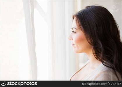 people and leisure concept - young woman looking through window at home. young woman looking through window at home