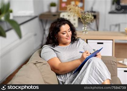 people and leisure concept - young woman with clipboard and pen taking notes on sofa at home. young woman with clipboard taking notes at home