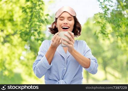 people and morning concept - happy young woman in pajama and eye sleeping mask drinking coffee from mug over green natural background. woman in pajama and sleeping mask drinking coffee