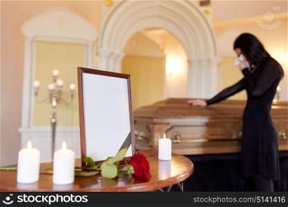 people and mourning concept - photo frame with black ribbon, candles and woman crying near coffin at funeral in church. photo frame and woman crying at coffin at funeral