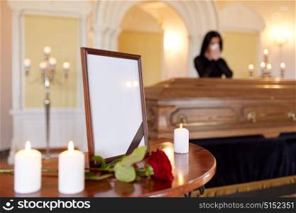 people and mourning concept - photo frame with black ribbon, candles and woman crying near coffin at funeral in church. photo frame and woman crying at coffin at funeral