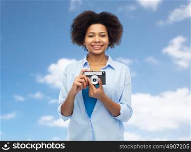 people and photography portrait concept - happy african american young woman with film camera over blue sky and clouds background. happy african american woman with film camera