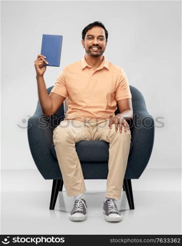people and reading concept - happy smiling young indian man sitting in chair showing book over grey background. happy young indian man showing book in chair