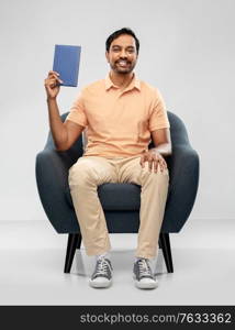 people and reading concept - happy smiling young indian man sitting in chair showing book over grey background. happy young indian man showing book in chair