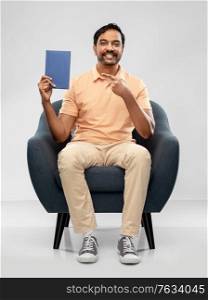 people and reading concept - happy smiling young indian man sitting in chair showing book over grey background. happy young indian man showing book in chair
