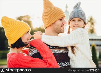 People and relationship concept. Family have unforgettable time together, embrae each other, wear trendy knitted hats. Adorable small girl with pigtails embrace her father, looks happily into camera